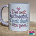 A white ceramic mug with a pastel message that reads 'I'm not antisocial, I just don't like you' in pink, purple, and blue bubble letters, with small heart and star accents. The mug is placed on a wooden surface against a neutral beige background.