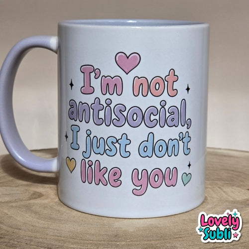 A white ceramic mug with a pastel message that reads 'I'm not antisocial, I just don't like you' in pink, purple, and blue bubble letters, with small heart and star accents. The mug is placed on a wooden surface against a neutral beige background.