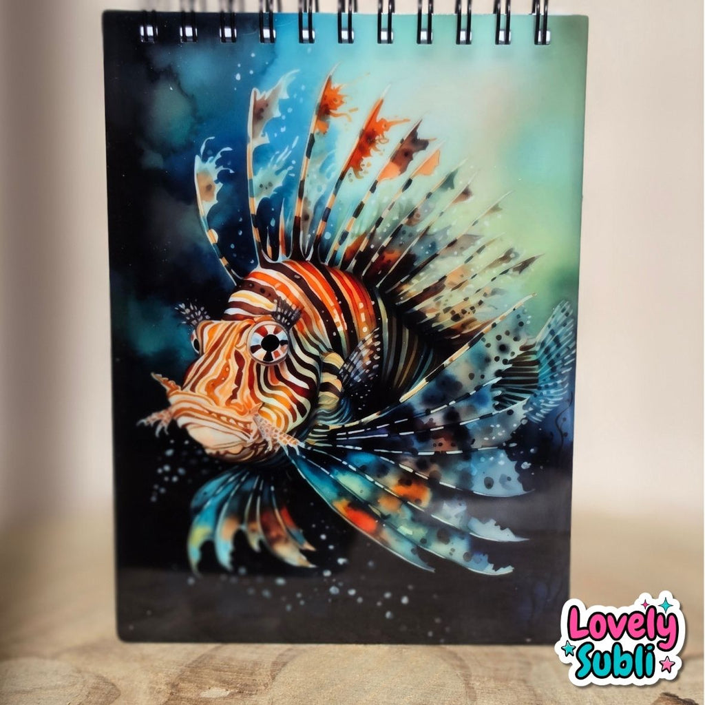 A spiral-bound notebook featuring a vibrant, detailed illustration of a lion fish on the cover, set against a dark underwater background. The notebook is standing upright on a wooden surface with a neutral beige background.