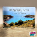 A rectangular souvenir A6 metal plaque featuring a scenic photograph of Lulworth Cove, Jurassic Coast, Dorset, with the text 'LULWORTH COVE' and coordinates printed on the front. The card is photographed standing upright on a sandy surface against a neutral beige background.