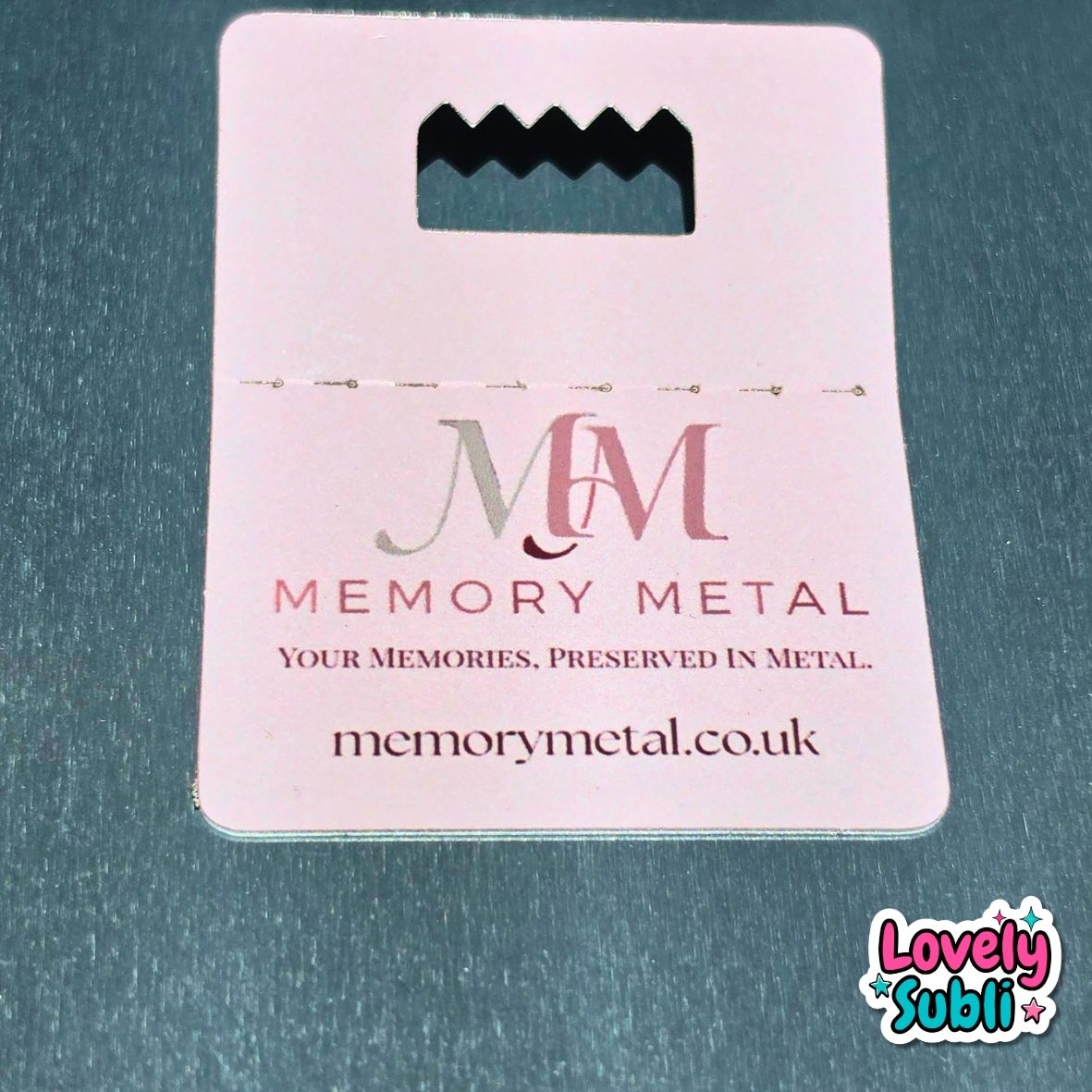 A pale pink Memory Metal keepsake card with a cutout at the top, printed with the Memory Metal logo and website, placed on a textured dark teal surface.