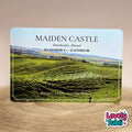 A rectangular A6 metal plaque featuring a landscape photo of green rolling hills under a pale blue sky, with the text 'MAIDEN CASTLE', coordinates, and 'Europe's Largest Iron-Are Hillfort'. The item sits on a wooden surface against a warm beige background.