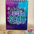 A spiral-bound notebook with a colorful geometric cover featuring the text 'Adulting IS A SCAM' in stylized lettering. The notebook is standing on a wooden surface against a softly blurred light background, with a 'Lovely Subli' watermark in the bottom right corner.