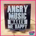 A square acrylic sign with the text 'ANGRY MUSIC MAKES ME HAPPY' in distressed white lettering on a black background, displayed on a small stand on a wooden surface.