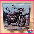 A square framed art print featuring a stylized illustration of a vintage motorcycle in a city street, displayed on a stand against a beige background with a pink border.