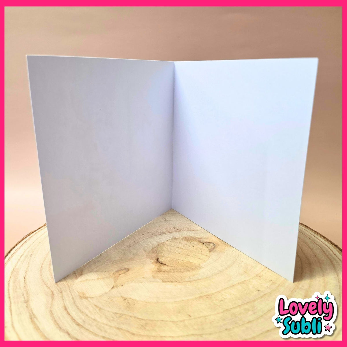 A blank white square card standing open in a V-shape or corner shape on a round wooden slice, set against a warm beige background with a pink border and a 'Lovely Subli' watermark in the bottom right corner.
