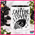 A white ceramic mug with a black handle and black interior, printed with the words 'CAFFEINE COVEN' and a skeletal hand holding a mug with moon and star details. The mug is photographed on a marble-like surface with a blurred dark background and greenery.