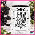 A white ceramic mug with a black handle and black interior, printed with black text reading 'I RUN ON CAFFEINE SARCASM & POOR DECISIONS' with moon and star graphics. The mug is photographed on a marble-like surface with blurred decorative background elements and greenery.