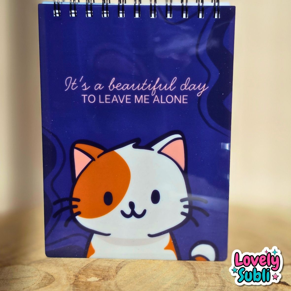 A spiral-bound notebook with a dark blue cover featuring a cartoon cat illustration and the text 'It's a beautiful day TO LEAVE ME ALONE'. The notebook is standing on a wooden surface against a warm beige background.