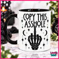 A white ceramic mug with a black handle and black interior, printed with the phrase 'COPY THIS ASSHOLE' above a black skeletal hand gesture and decorative stars and moons. The mug is photographed on a marble-like surface with a blurred indoor background and a pink border around the image.