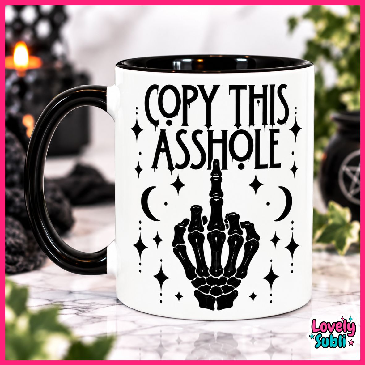 A white ceramic mug with a black handle and black interior, printed with the phrase 'COPY THIS ASSHOLE' above a black skeletal hand gesture and decorative stars and moons. The mug is photographed on a marble-like surface with a blurred indoor background and a pink border around the image.