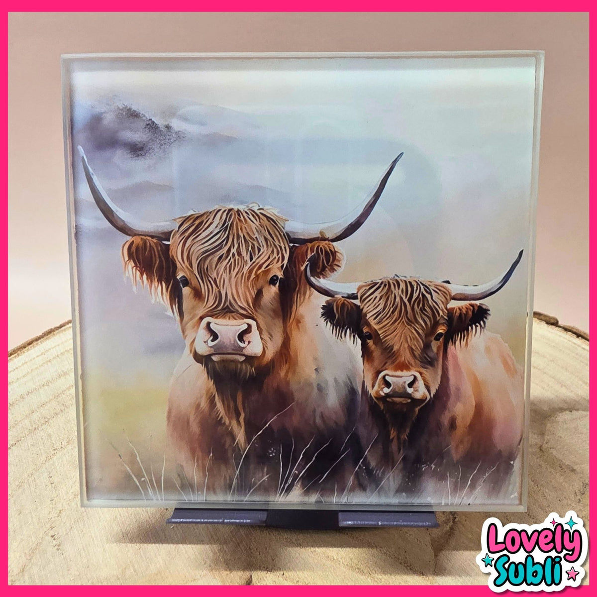 A square acrylic art plaque featuring a painted portrait of two Highland cows, displayed on a small stand on a wooden surface. The plaque is framed in clear acrylic and has a pink border.