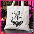 A white canvas tote bag with black printed text reading 'CUTE BUT CREEPY' above a black moth with a skull in the center, displayed on a dark table with gothic-themed props including a skull, candle, and books.