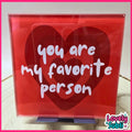 A square acrylic sign with a red background featuring three overlapping heart shapes and the phrase 'you are my favorite person' in white script. The sign is displayed on a small stand on a light surface, with a 'Lovely Subli' watermark in the bottom right corner.