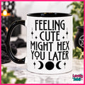 A white ceramic mug with a black handle and black interior, printed with the phrase 'FEELING CUTE MIGHT HEX YOU LATER' in black gothic-style lettering, alongside moon phases. The mug is shown on a marble surface with a blurred Halloween-themed background.