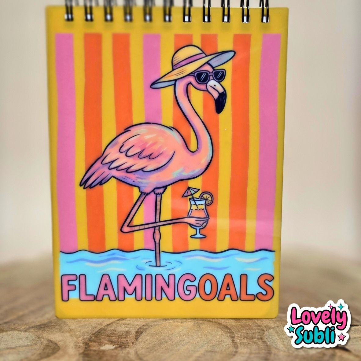 A spiral-bound notebook featuring a colorful flamingo illustration on the cover, standing on a wooden surface against a light beige background. The cover has vertical pink and orange stripes, a pink flamingo wearing a yellow hat and sunglasses, a cocktail glass, and the text 'FLAMINGOALS' at the bottom.