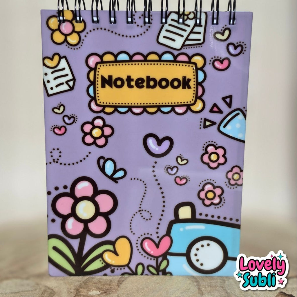 A spiral-bound notebook with a lavender cover decorated with colorful doodle-style flowers, hearts, butterflies, and a blue car. The center features a scalloped yellow label with the word 'Notebook' in black.