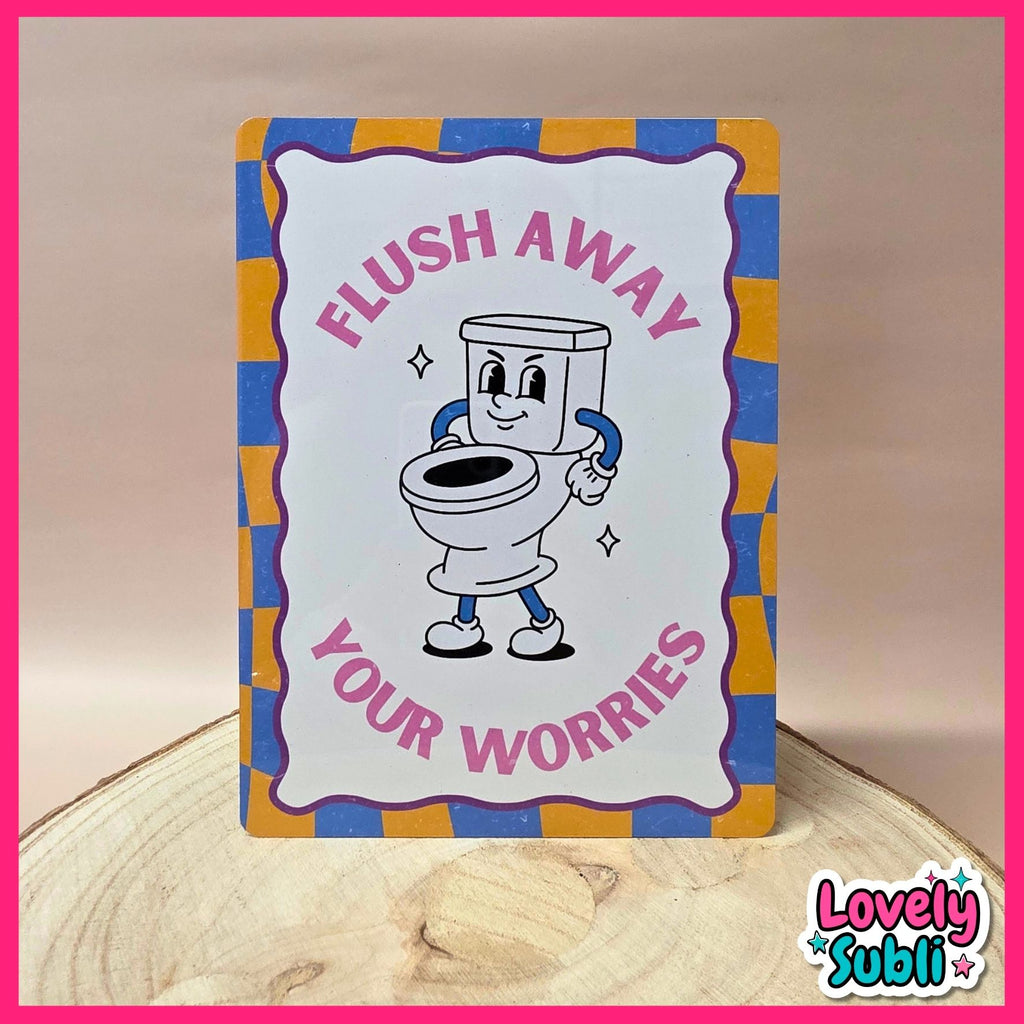 A rectangular novelty sign with a cartoon toilet character and the text 'FLUSH AWAY YOUR WORRIES,' photographed standing on a wood slice against a beige background. The sign has a white center, pink lettering, and a blue-and-yellow checkered border.