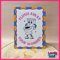 A rectangular novelty sign with a cartoon toilet character and the text 'FLUSH AWAY YOUR WORRIES,' photographed standing on a wood slice against a beige background. The sign has a white center, pink lettering, and a blue-and-yellow checkered border.