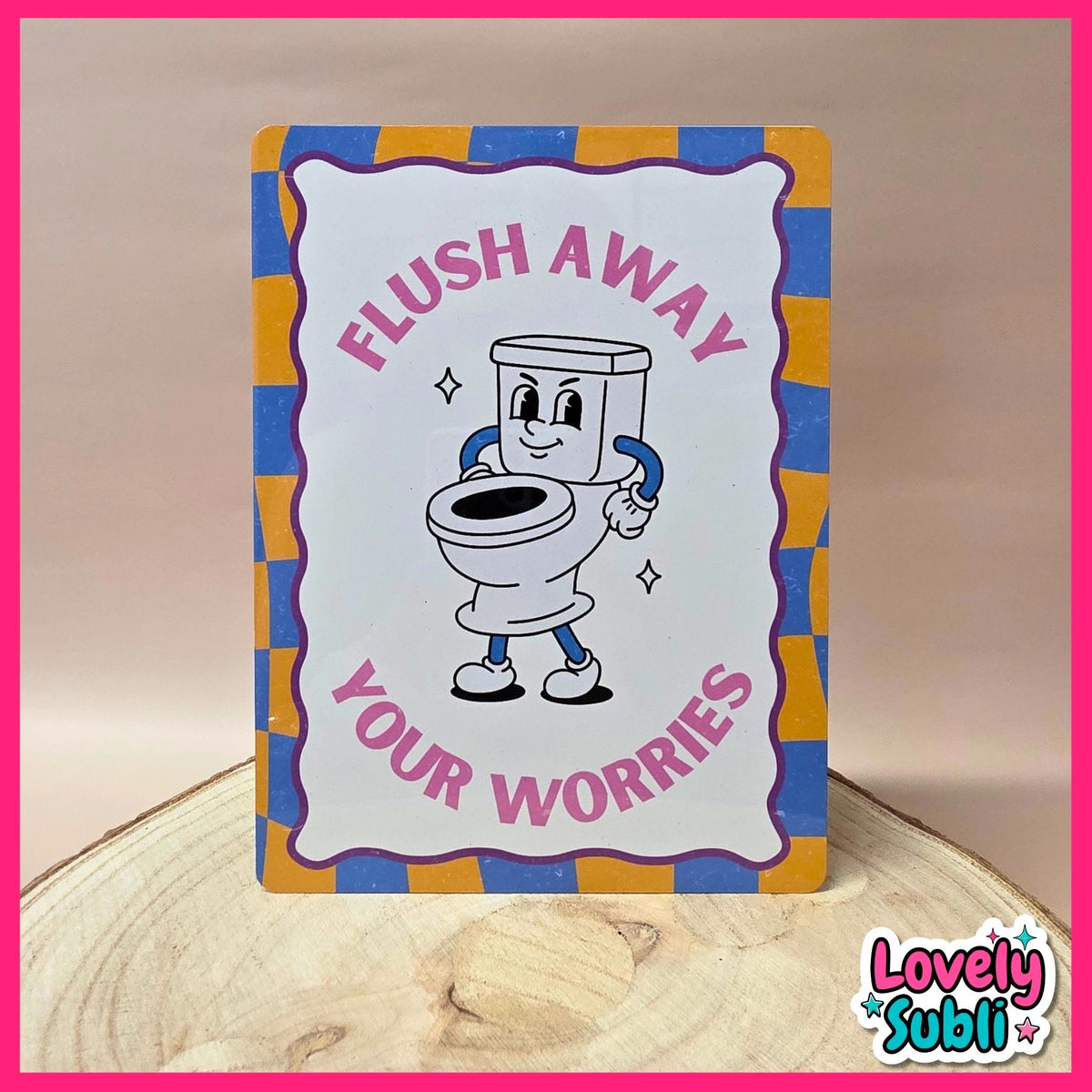 A rectangular novelty sign with a cartoon toilet character and the text 'FLUSH AWAY YOUR WORRIES,' photographed standing on a wood slice against a beige background. The sign has a white center, pink lettering, and a blue-and-yellow checkered border.