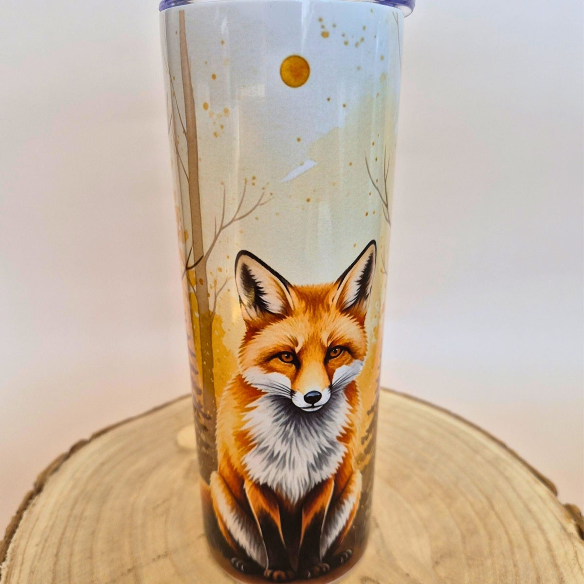 A tall insulated tumbler with a lid, featuring a detailed illustration of a red fox in a light-colored forest scene, displayed on a round wooden slice against a plain light background.