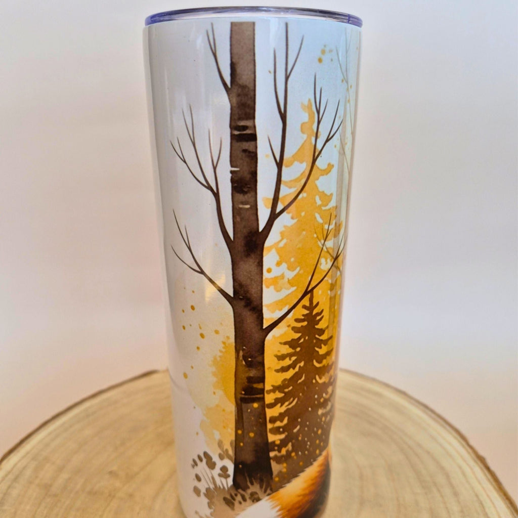 A tall insulated tumbler with a clear winter forest design, featuring a birch tree, pine trees, and a fox, displayed on a round wooden slice against a neutral background.