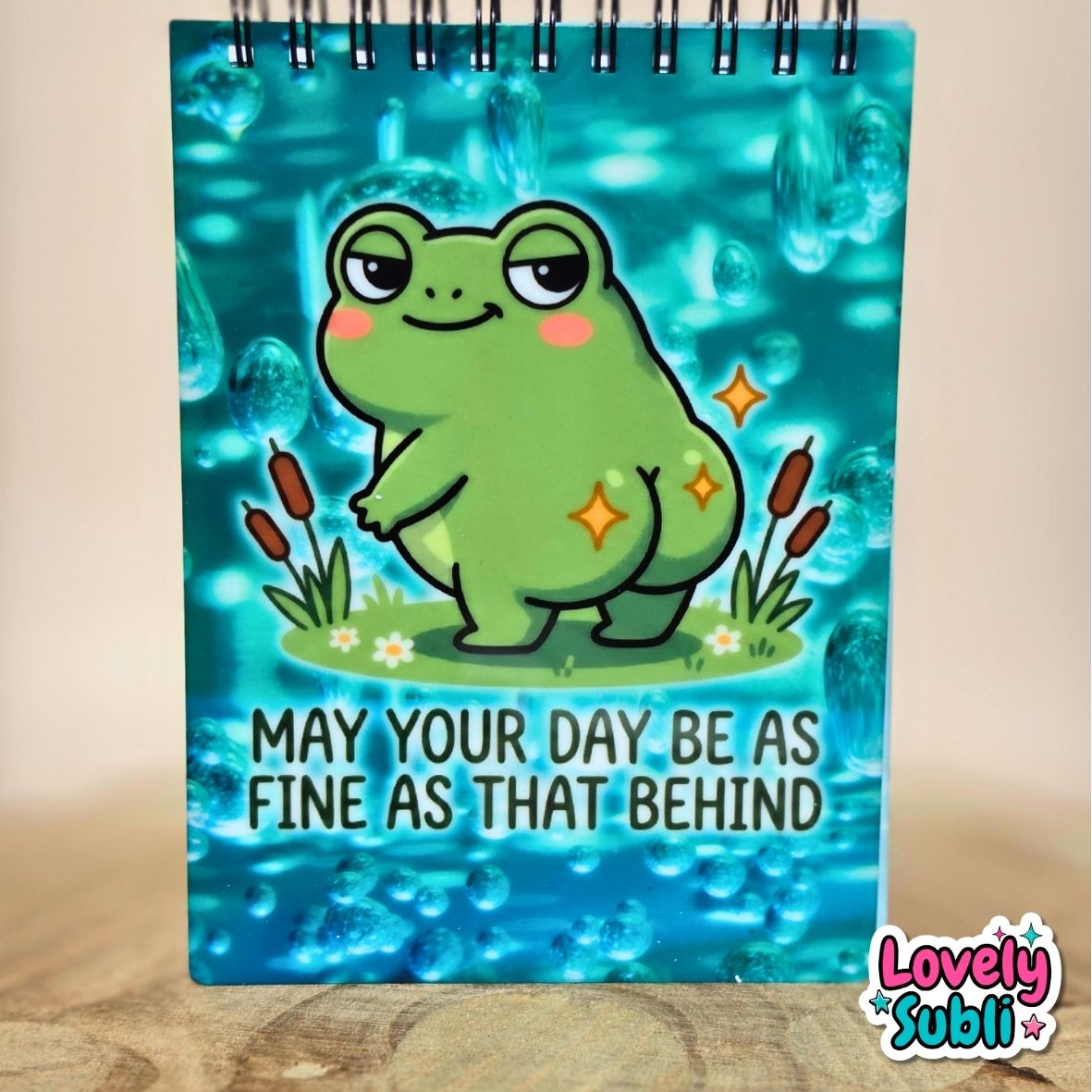 A spiral-bound notebook with a cartoon frog illustration on a blue water-themed background. The text on the cover reads 'MAY YOUR DAY BE AS FINE AS THAT BEHIND'.