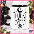 A white ceramic mug with a black handle and black interior, featuring a black graphic print of a dripping crescent moon, stars, vertical text reading 'FUCK OFF', and a pentagram. The mug is photographed on a marble-like surface with a blurred gothic-themed background.