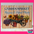 A rectangular decorative sign with a rustic wood-plank background and the text 'GARDEN MARKET Flowers & Potted Plants.' The sign features an illustrated wheelbarrow filled with sunflowers, red flowers, green plants, purple flowers, yellow flowers, a copper watering can, butterflies, and potted plants.