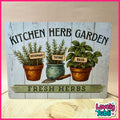 A decorative rectangular sign reading 'KITCHEN HERB GARDEN' and 'FRESH HERBS,' featuring illustrated rosemary, thyme, and basil plants in pots with small gardening tools. The sign is photographed on a light wood surface against a beige wall, with a pink border around the image and a 'Lovely Subli' logo in the lower right corner.