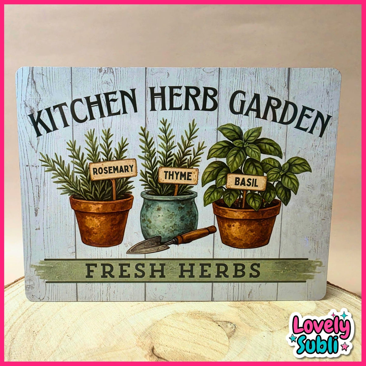 A decorative rectangular sign reading 'KITCHEN HERB GARDEN' and 'FRESH HERBS,' featuring illustrated rosemary, thyme, and basil plants in pots with small gardening tools. The sign is photographed on a light wood surface against a beige wall, with a pink border around the image and a 'Lovely Subli' logo in the lower right corner.