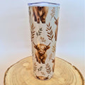 A tall insulated tumbler with a clear lid, decorated with a pattern of Highland cows and brown leaves on a cream background, displayed on a round wood slice against a light gray background.