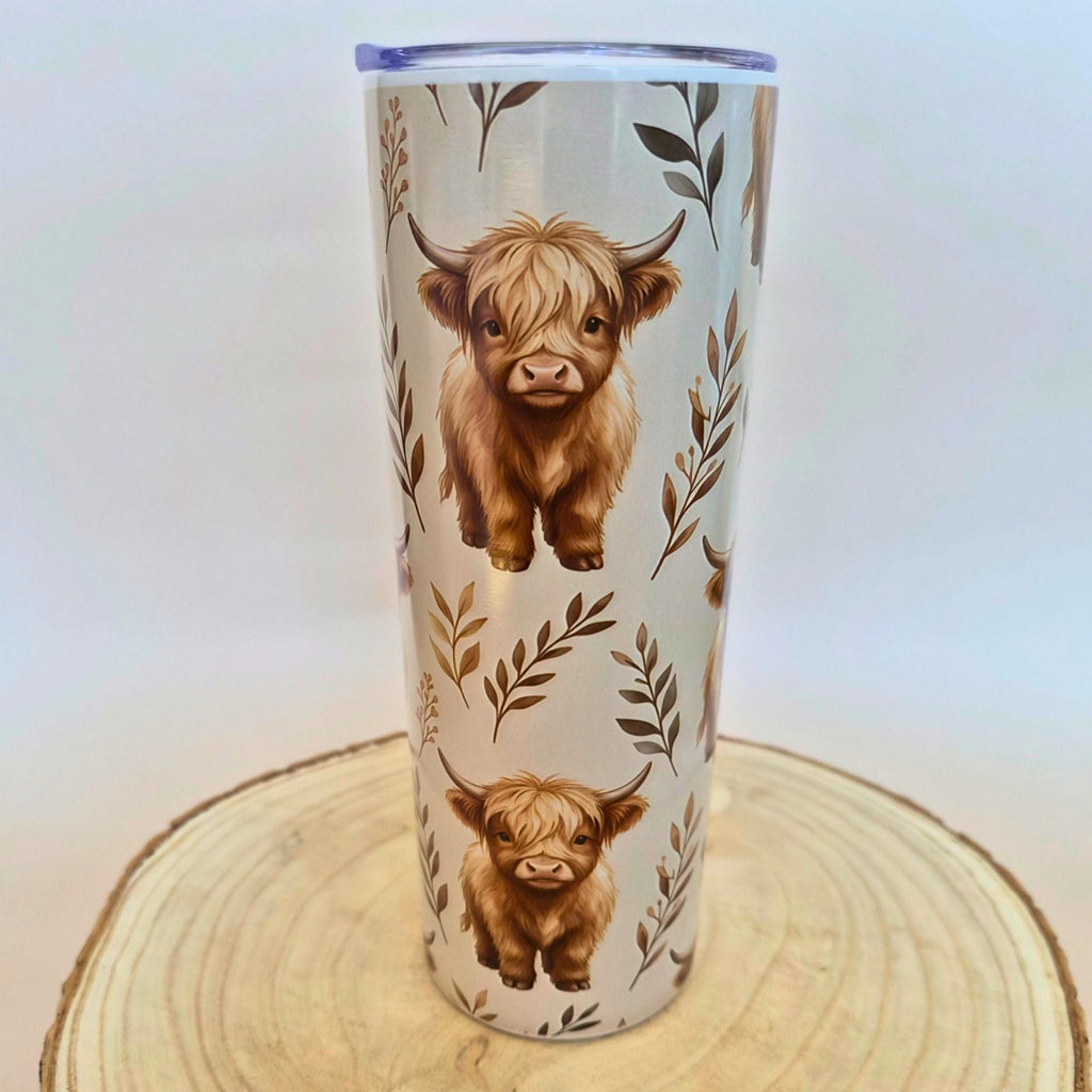 A tall insulated tumbler with a clear lid, decorated with two brown Highland cow illustrations and leafy botanical motifs on a light background, displayed on a round wood slice against a plain light backdrop.