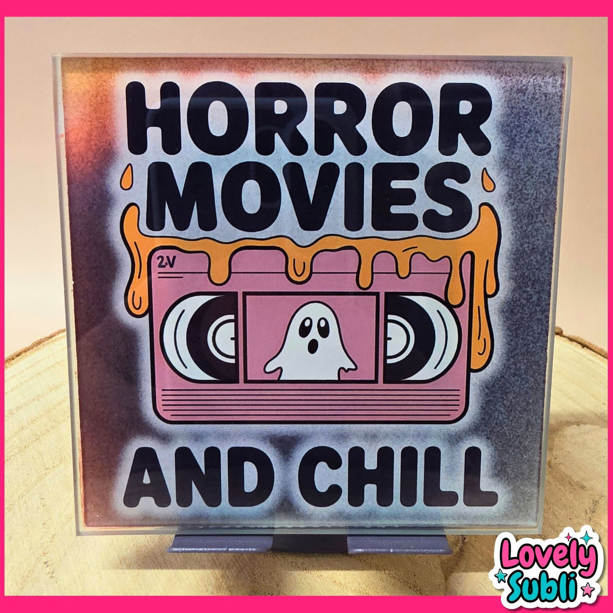 A square decorative sign or print featuring the phrase 'HORROR MOVIES AND CHILL' in bold black letters around a retro pink VHS tape illustration with a white ghost. The sign is displayed on a small stand on a wooden surface, with a pink border framing the image.