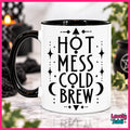 A white ceramic mug with a black handle and black interior, featuring the text 'HOT MESS COLD BREW' in black lettering with decorative symbols. The mug is set against a softly blurred background with a pink border.