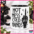 A white ceramic mug with a black handle and black interior, printed with the phrase 'HOT MESS COLD HANDS' in black lettering alongside a black skeletal hand graphic. The mug is photographed on a reflective surface with blurred decorative background elements, framed by a pink border.