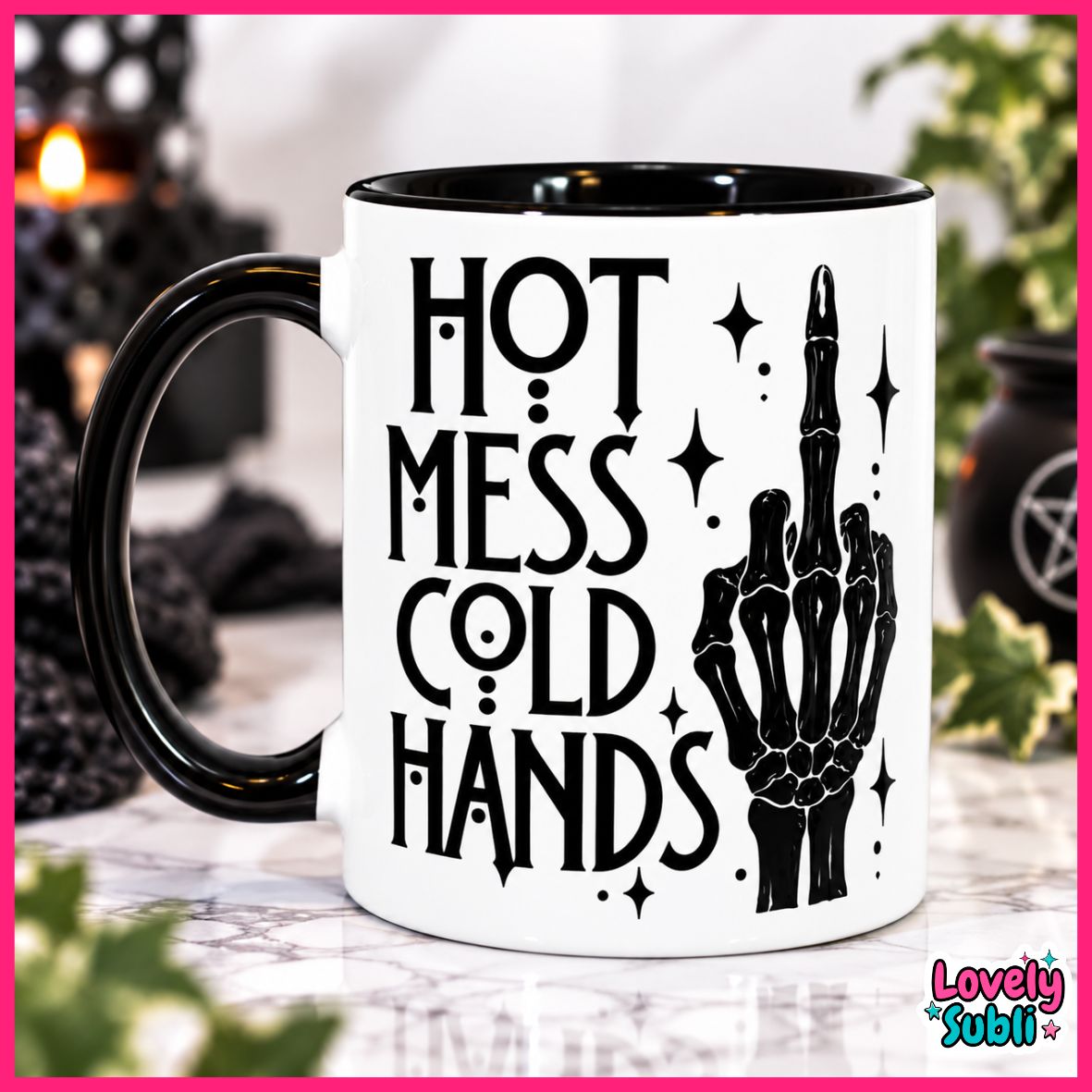 A white ceramic mug with a black handle and black interior, printed with the phrase 'HOT MESS COLD HANDS' in black lettering alongside a black skeletal hand graphic. The mug is photographed on a reflective surface with blurred decorative background elements, framed by a pink border.