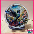 A round glass art piece featuring a detailed painted hummingbird with pink flowers and a stained-glass style border, displayed on a small stand. The background is a soft beige studio setting with a wood slice prop, framed by a pink border.