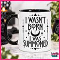 A white ceramic mug with a black handle and black interior, printed with the phrase 'I WASN'T BORN I WAS SUMMONED' in black gothic-style lettering with a crescent moon and star accents. The mug is shown on a marble-like surface with a blurred mystical background and a pink border around the image.