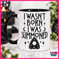 A white ceramic mug with a black handle and black interior, printed with the phrase 'I WASN'T BORN I WAS SUMMONED' in black lettering above a black triangular occult-style symbol. The mug is photographed on a marble-like surface with blurred black candles and greenery in the background.
