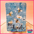 A rectangular decorative sign or card with rounded corners, featuring a blue watercolor-style background with illustrated bees, white daisies, and the words “BEE kind” in black script. The sign is displayed upright on a small stand on a wooden surface against a peach-pink backdrop.