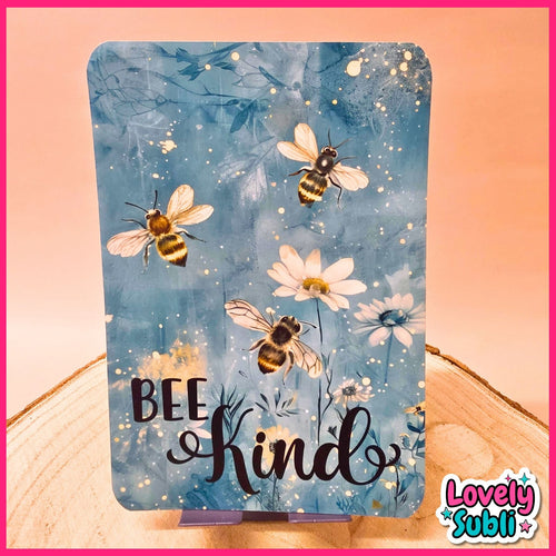 A rectangular decorative sign or card with rounded corners, featuring a blue watercolor-style background with illustrated bees, white daisies, and the words “BEE kind” in black script. The sign is displayed upright on a small stand on a wooden surface against a peach-pink backdrop.