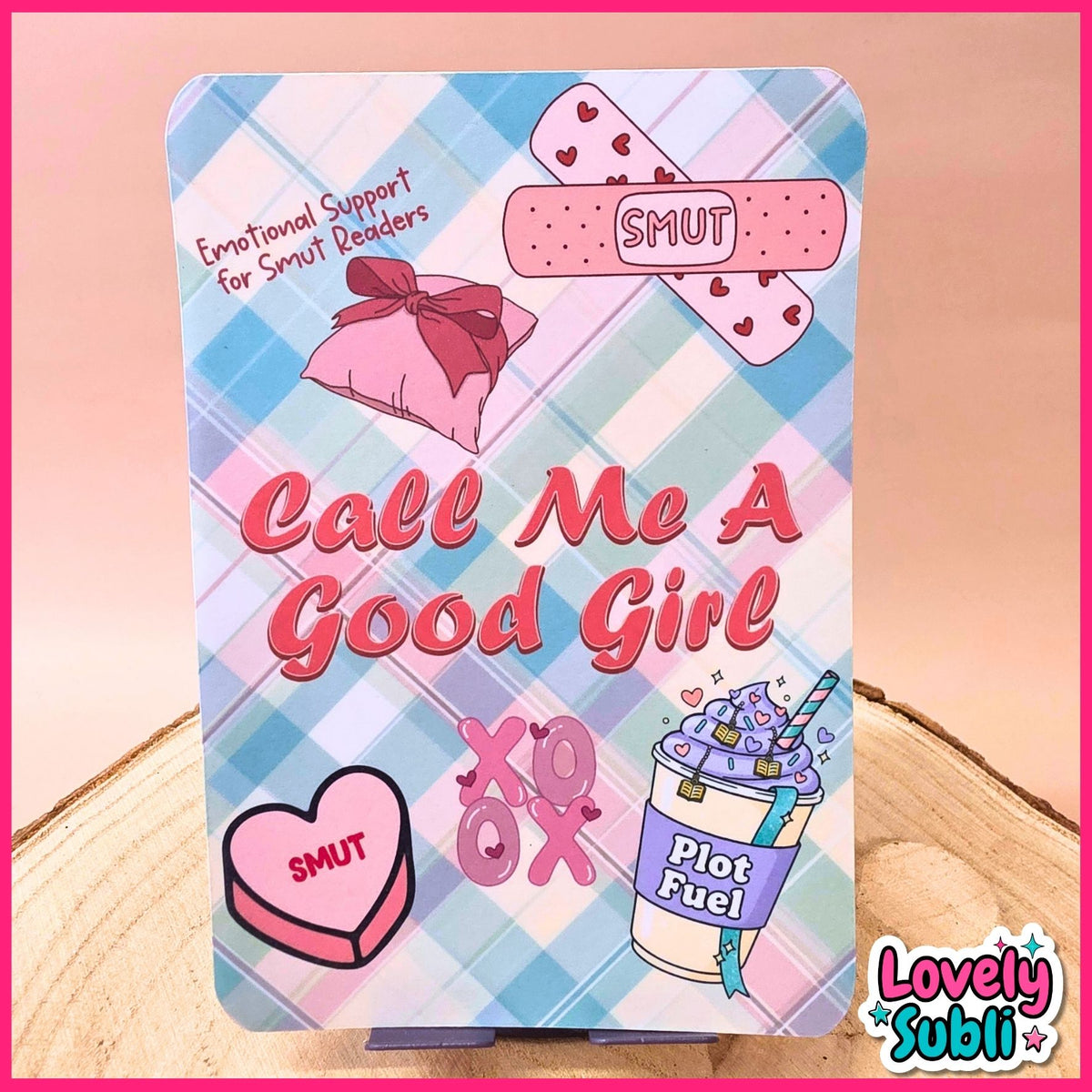 A square decorative sign or plaque with rounded corners featuring a pastel plaid background and cute pink-themed illustrations, including a band-aid with hearts, a pink gift box, a heart-shaped candy labeled 'SMUT', 'Call Me A Good Girl' in large pink text, and a drink cup labeled 'Plot Fuel'. The sign is displayed on a small stand on a wooden surface with a pink backdrop.
