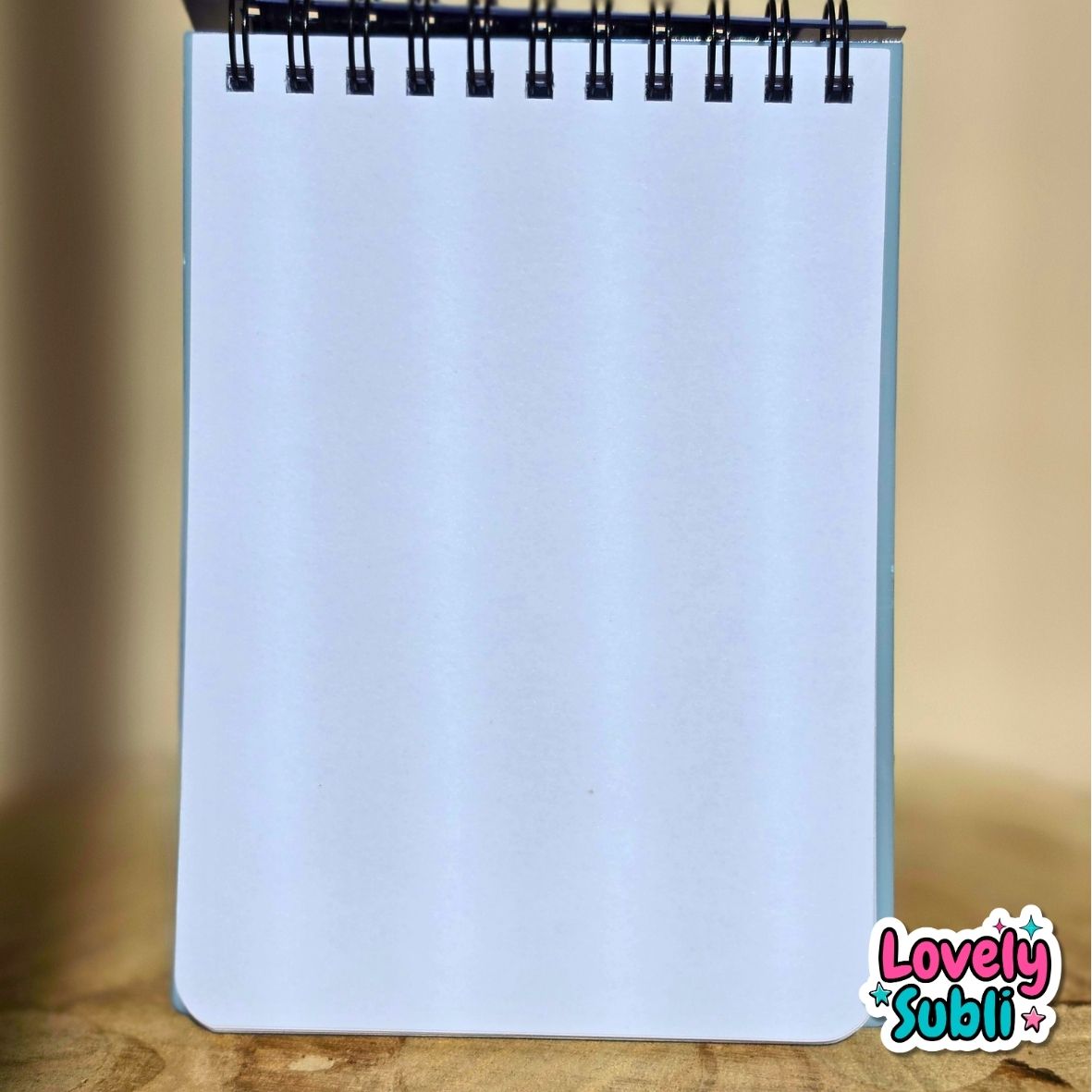 A spiral-bound notepad with blank white pages standing upright on a wooden surface, photographed against a softly blurred beige background. A 'Lovely Subli' brand watermark is visible in the bottom right corner.