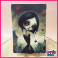 A vertical art print or card featuring a gothic illustration of a girl with large dark eyes, black hair, and a black dress, surrounded by red hearts in a misty village scene. The print is displayed upright on a wooden surface against a pale pink background with a decorative pink border and a 'Lovely Subli' logo in the bottom right corner.