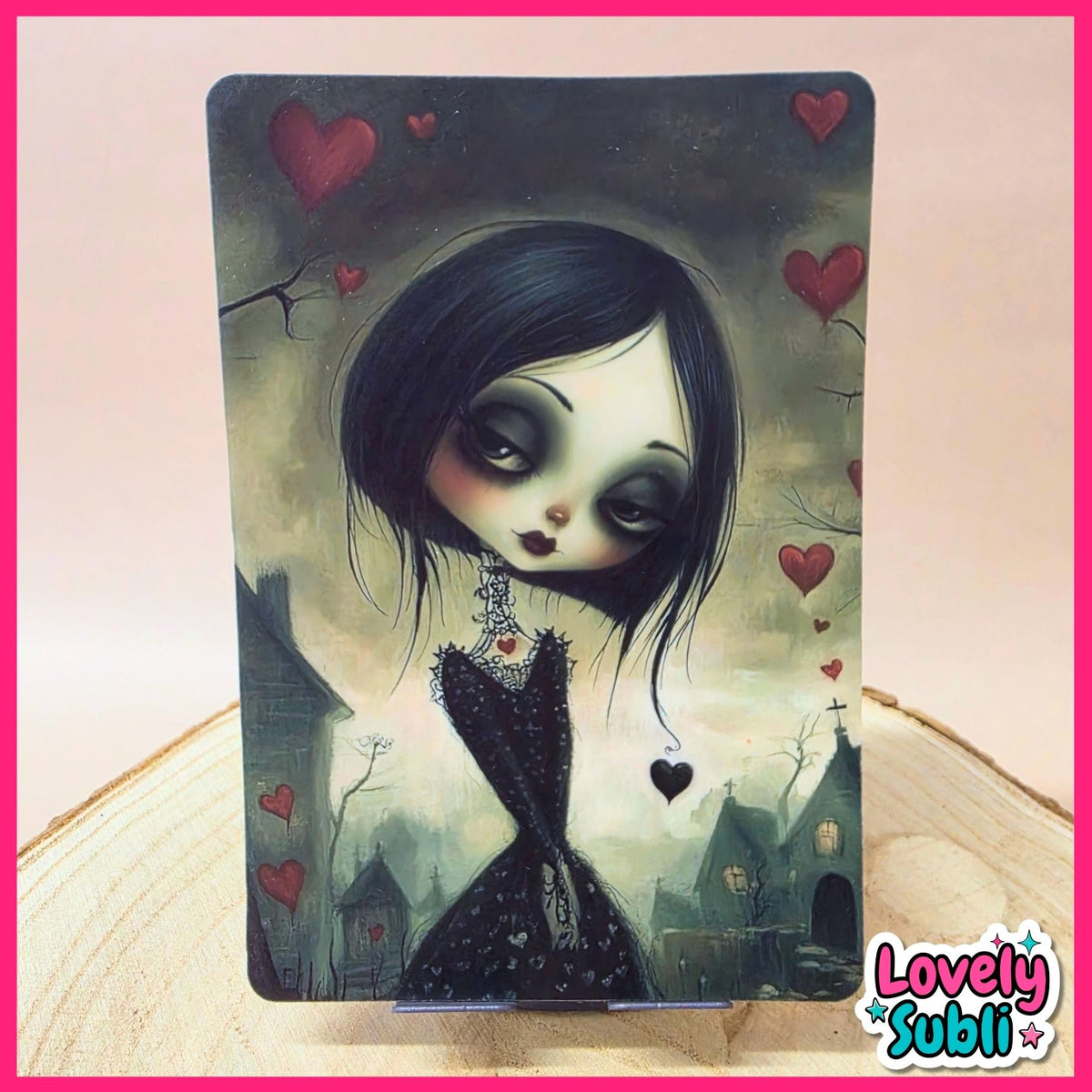 A vertical art print or card featuring a gothic illustration of a girl with large dark eyes, black hair, and a black dress, surrounded by red hearts in a misty village scene. The print is displayed upright on a wooden surface against a pale pink background with a decorative pink border and a 'Lovely Subli' logo in the bottom right corner.