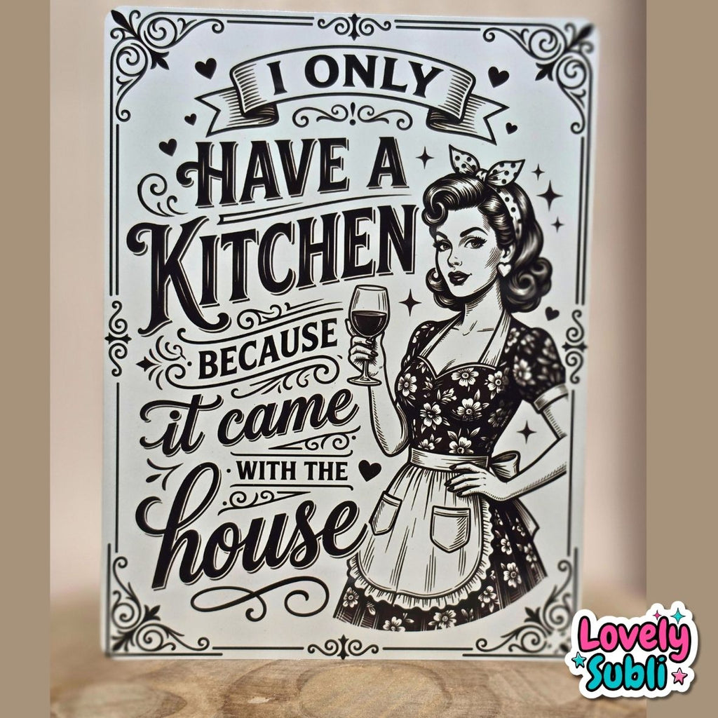 A rectangular decorative sign with a vintage-style black and white illustration of a woman in a floral dress holding a wine glass, surrounded by decorative text reading 'I ONLY HAVE A KITCHEN BECAUSE it came with the house.' The sign is photographed standing upright on a wooden surface against a beige wall.