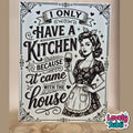 A rectangular decorative sign with a vintage-style black and white illustration of a woman in a floral dress holding a wine glass, surrounded by decorative text reading 'I ONLY HAVE A KITCHEN BECAUSE it came with the house.' The sign is photographed standing upright on a wooden surface against a beige wall.