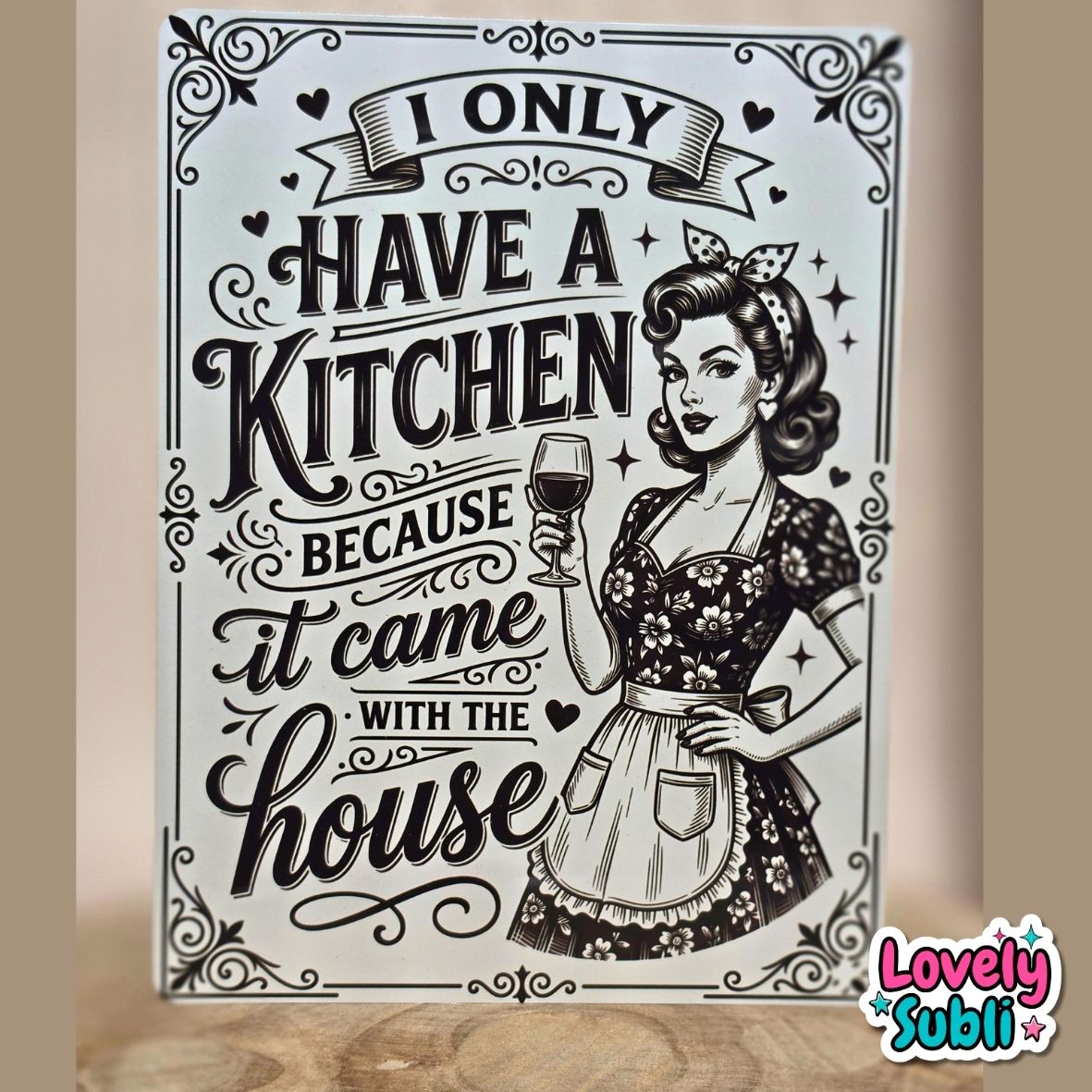 A rectangular decorative sign with a vintage-style black and white illustration of a woman in a floral dress holding a wine glass, surrounded by decorative text reading 'I ONLY HAVE A KITCHEN BECAUSE it came with the house.' The sign is photographed standing upright on a wooden surface against a beige wall.