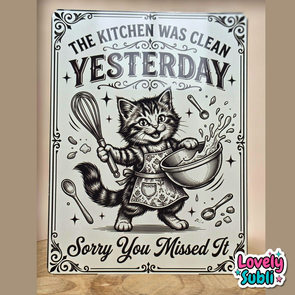A rectangular decorative sign with a vintage-style black-and-white illustration of a cat in an apron holding a whisk and mixing bowl, with the text 'THE KITCHEN WAS CLEAN YESTERDAY Sorry You Missed It.' The sign is photographed standing upright against a beige wall on a wooden surface.